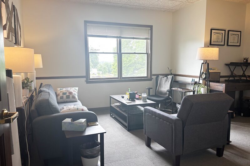A bright counseling room with a sofa, armchair, and a large window